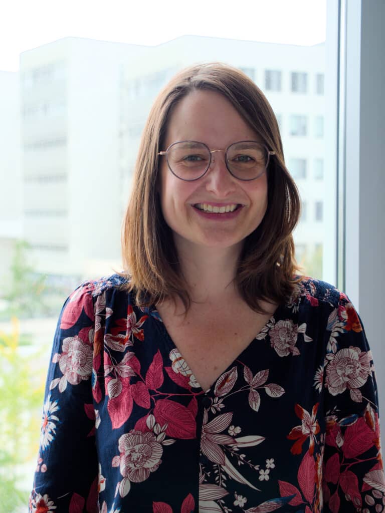 Photographie de Marianne Vigneulle. Cheveux châtains au carré lui tombant sur les épaules, lunettes rondes, elle porte une chemise bleu nuit à motifs fleuris roses et ocre. Elle sourit en regardant l'objectif. Derrière elle, une fenêtre donne sur les bâtiments blancs et les espaces verts du campus Condorcet d’Aubervilliers, où sont situés les bureaux du PPR.