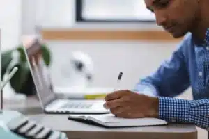 Main d'un homme qui écrit sur un bloc note, devant un ordinateur.
