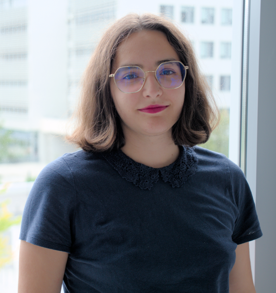 Photographie de Marie Assaf. Les cheveux bruns coupés au carré, elle porte des lunettes rondes discrètes et un t-shirt bleu nuit. Elle sourit à la caméra. En arrière-plan apparaissent les immeubles blancs du campus Condorcet d'Aubervilliers.