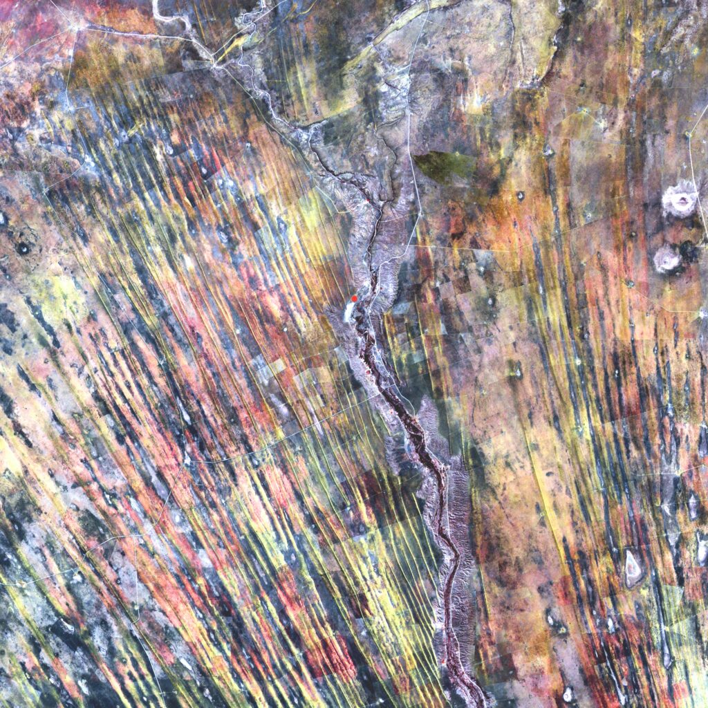 Terre vue du ciel : roches grises striées avec des reflets irisés. Une route serpente au centre de la photographie et se divise dans la partie supérieure.