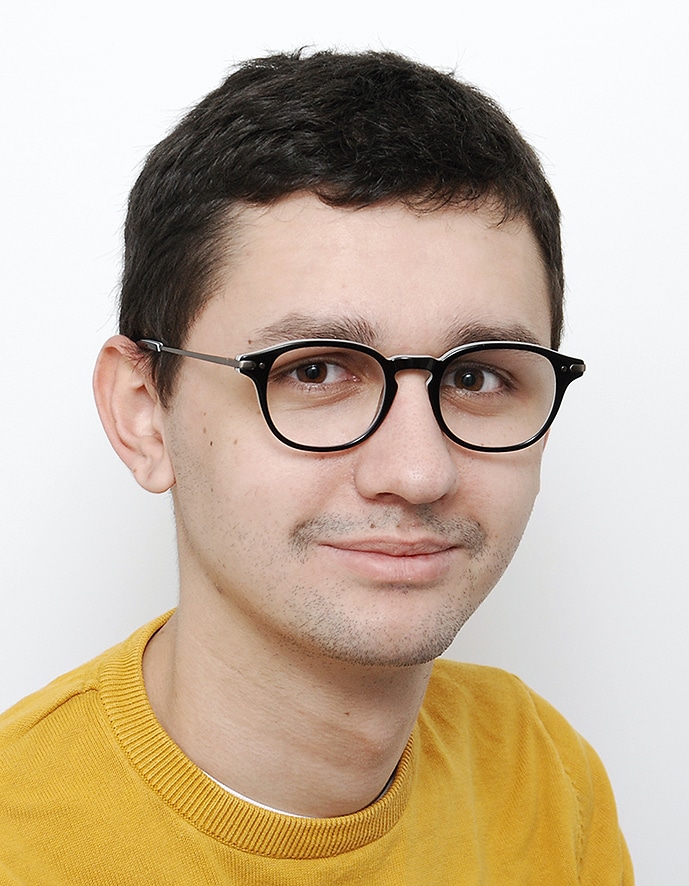 Photographie de Léandre Guignier. Cheveux bruns courts, lunettes noires et t-shirt jaune, il sourit à l’objectif.