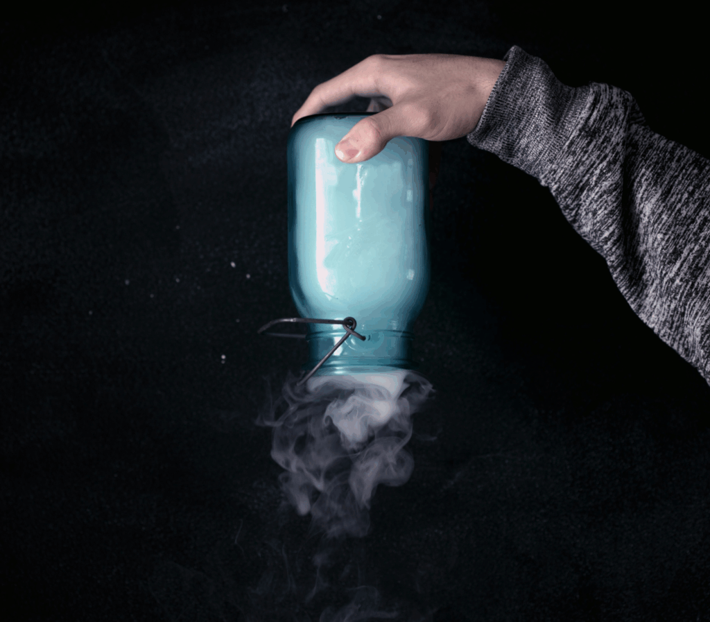 Photographie d'une main tenant un bocal en verre de couleur bleu-vert. L'ouverture est dirigée vers le bas, de la fumée blanche s'en échappe.